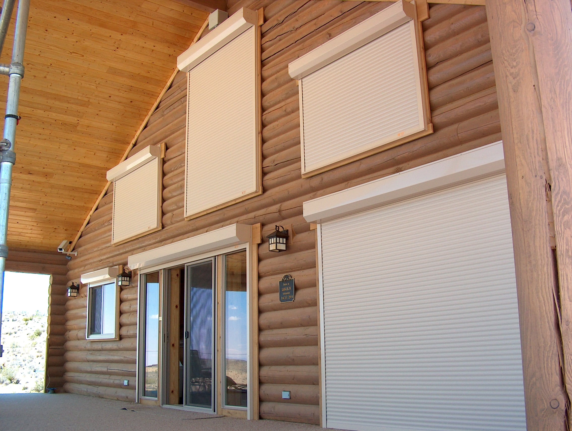 Two-story stucco home with multiple Roll-A-Shield rolling shutters installed across upper and lower windows and a patio door