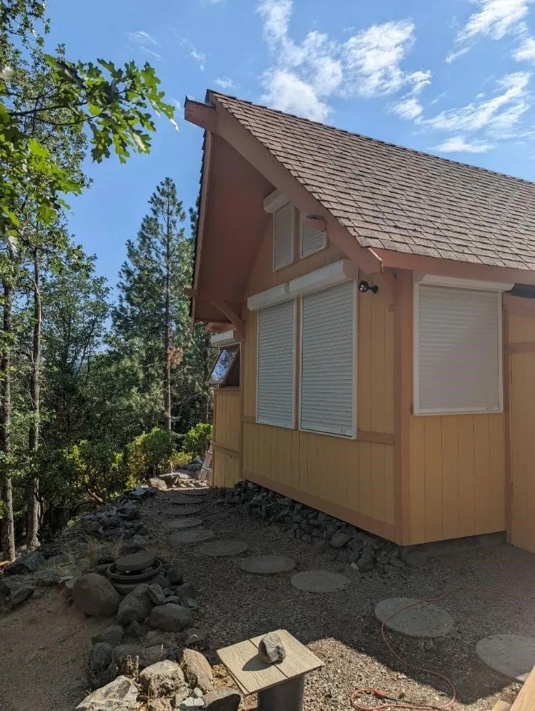 Wood-sided cabin in a wildfire-prone setting with white rolling shutters protecting the windows