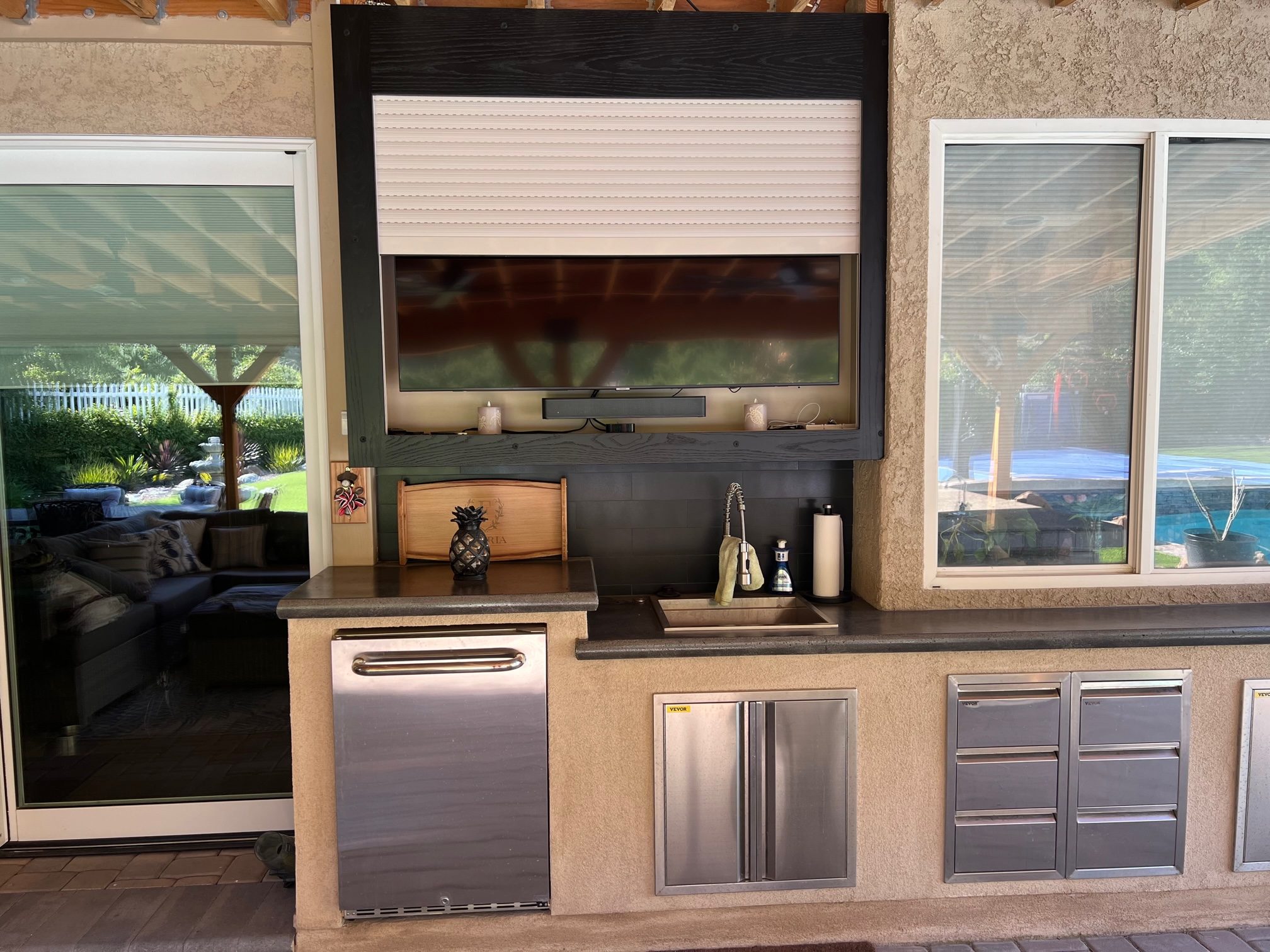 White rolling shutter partly lowered over an outdoor television above an outdoor kitchen with a sink and refrigerator
