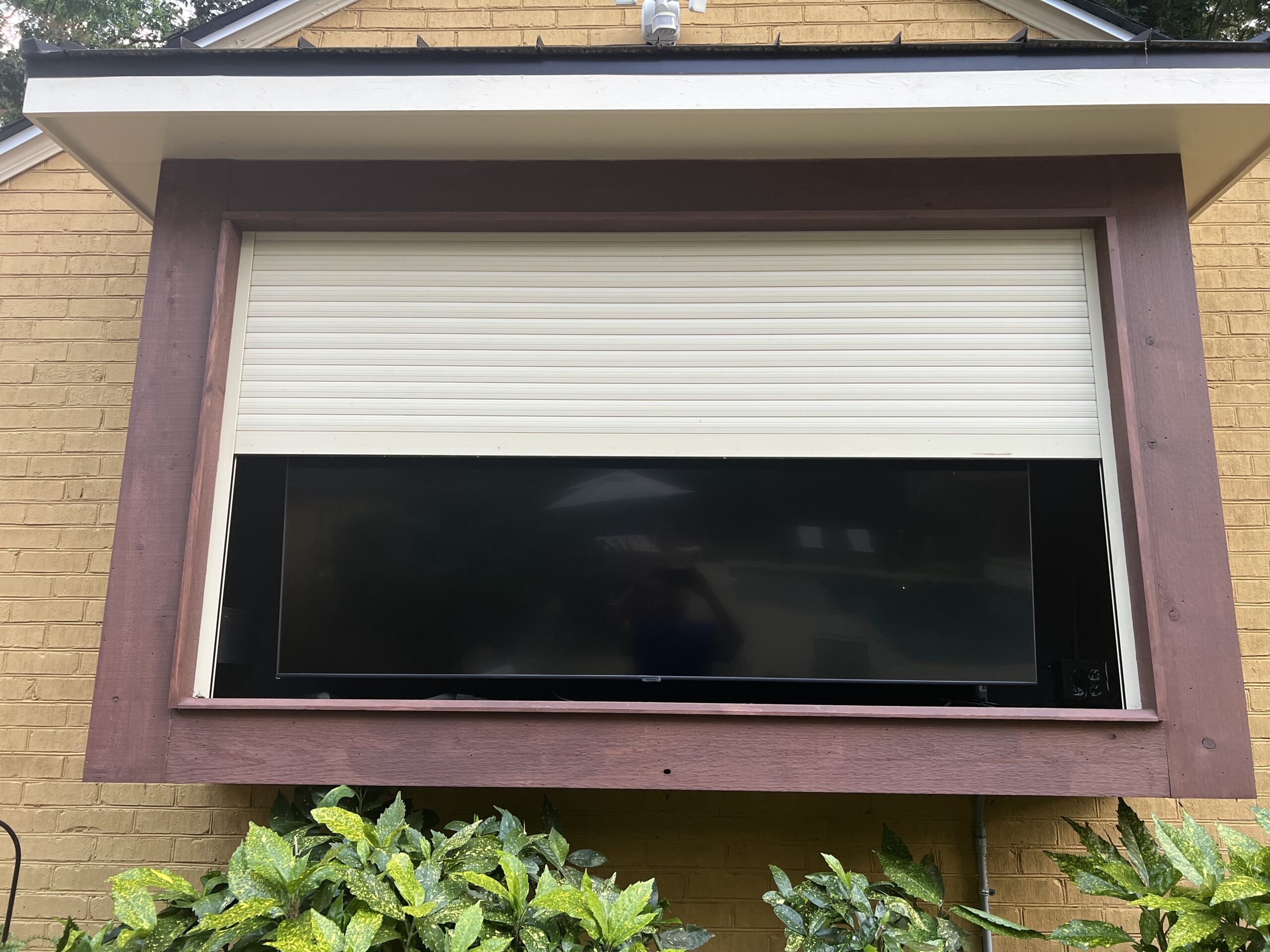 Cream rolling shutter half lowered over a wide outdoor television recessed in a timber-framed opening in a brick wall