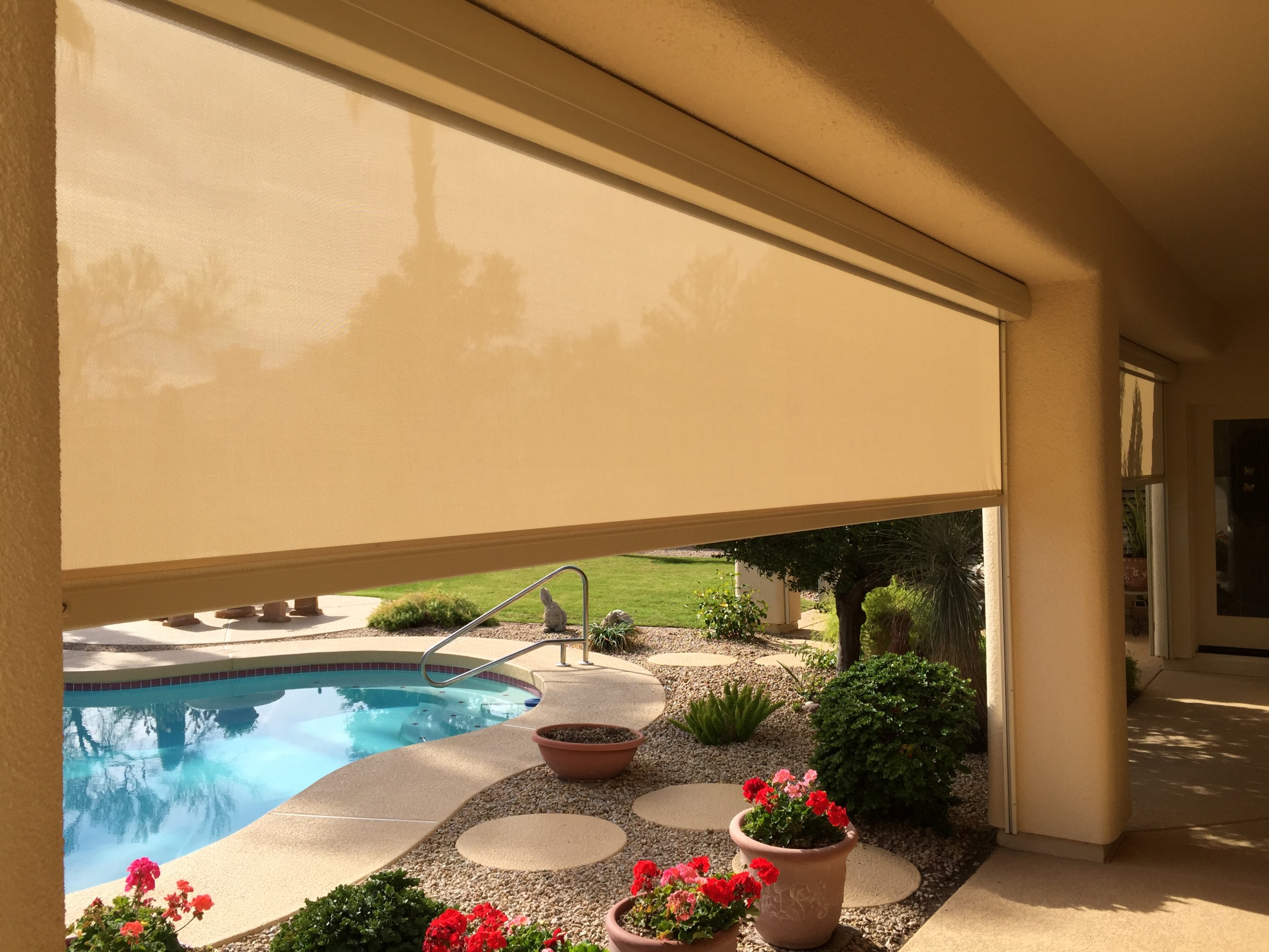Sand-coloured motorized rolling screen part way down across a covered patio opening, with the pool and yard visible below it