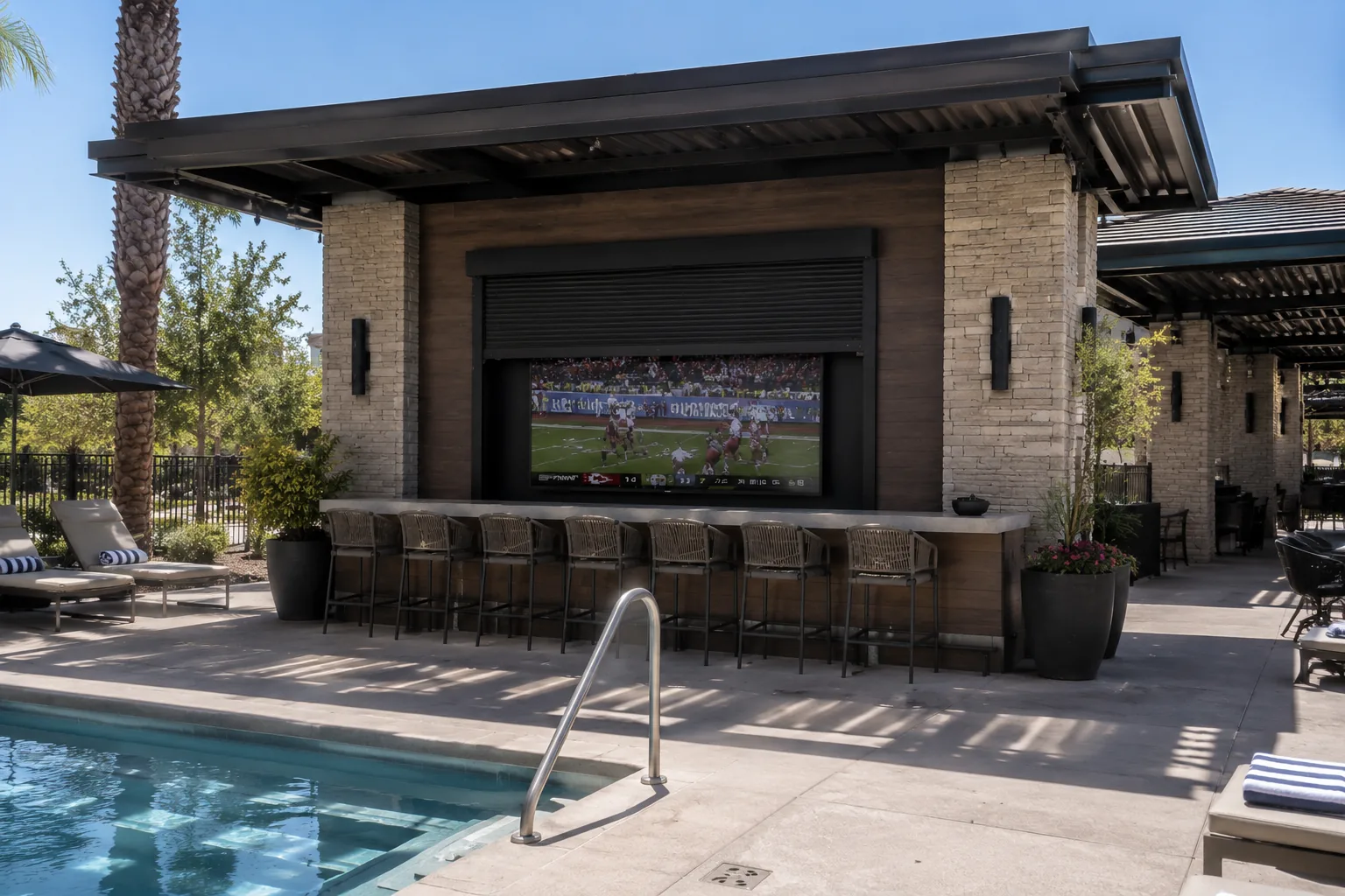 Black rolling shutter raised above a large outdoor television at a poolside bar with stool seating