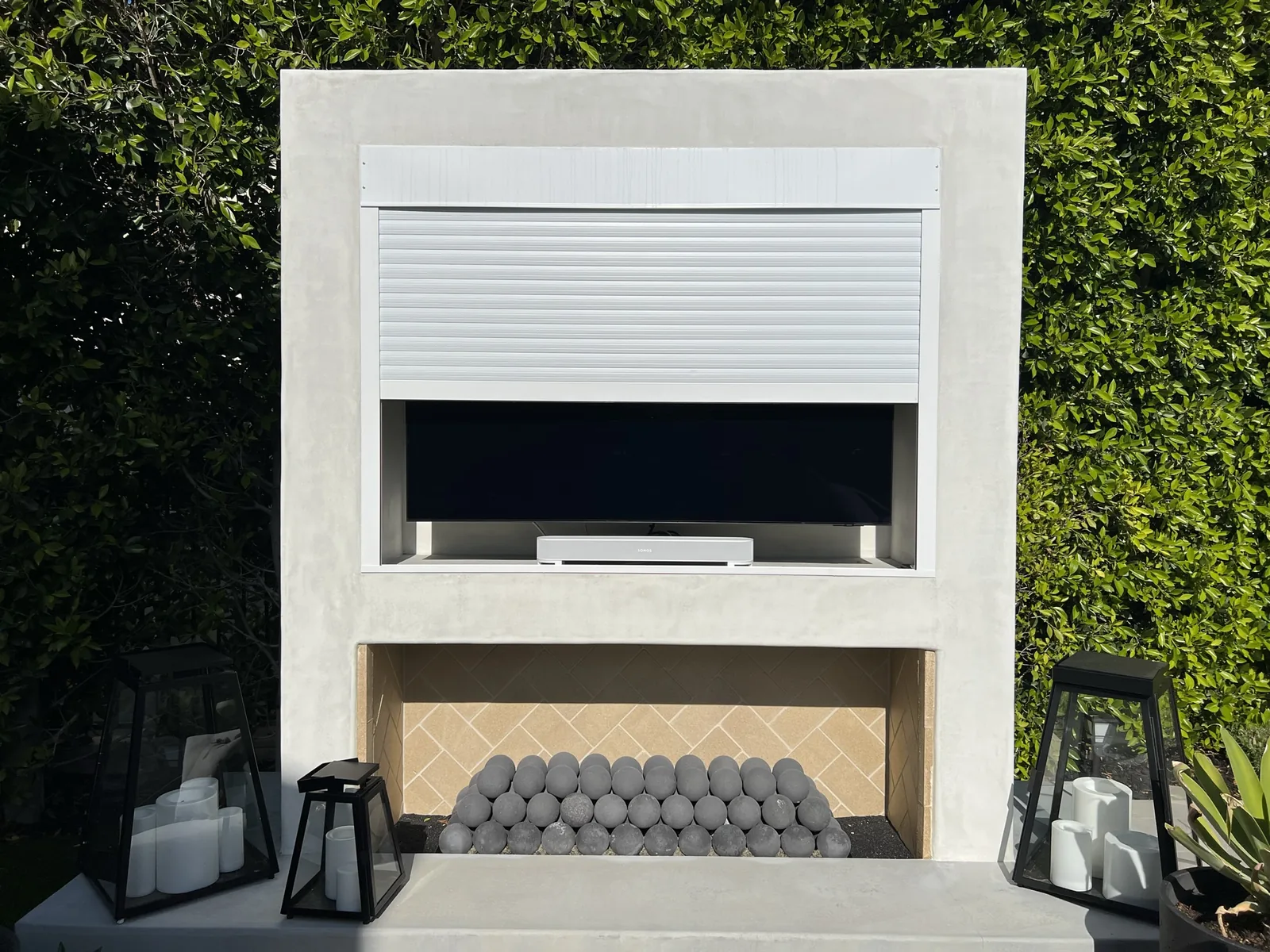 White rolling shutter half closed over a wall-mounted outdoor television set into a stucco media wall above a gas fire feature