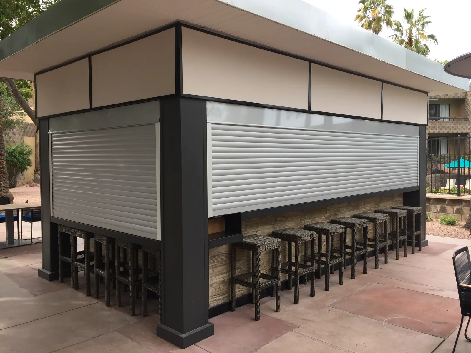 Rolling shutters closed over the service counters of an outdoor poolside bar, with bar stools lined up below