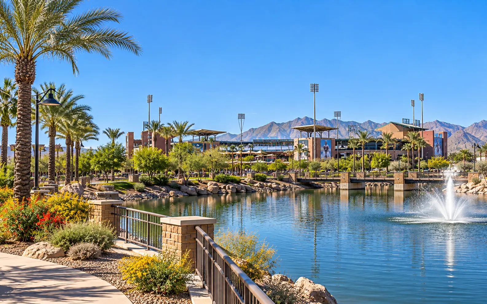 Lakeside walking path in Goodyear, Arizona, with palm trees, desert landscaping, and a fountain, and a ballpark and mountains beyond