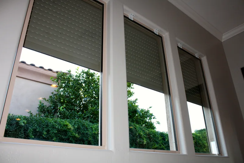 Rolling shutters lowered partway over a row of windows, seen from inside a Peoria home, with greenery visible through the open lower portion