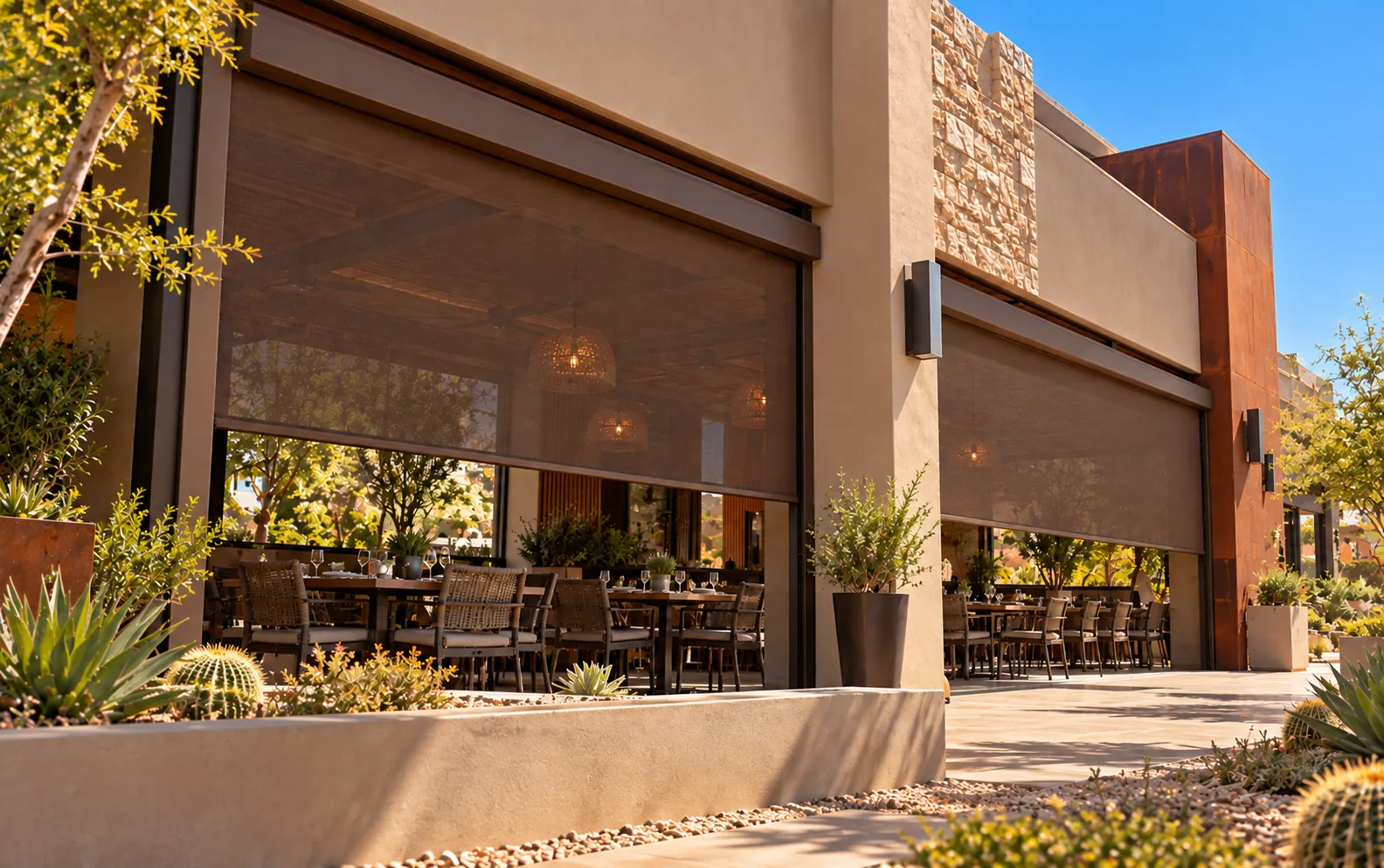 Motorized screens partly lowered across the open front of a covered restaurant patio on a sunny day, shading set dining tables behind a desert garden of cactus and agave