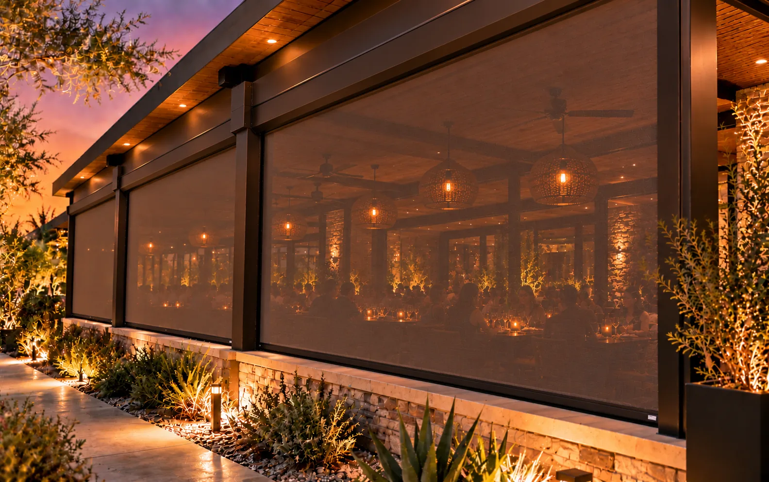 Motorized screens fully lowered around a covered restaurant patio at dusk, with the lit dining area glowing through the mesh and landscape lighting along the path