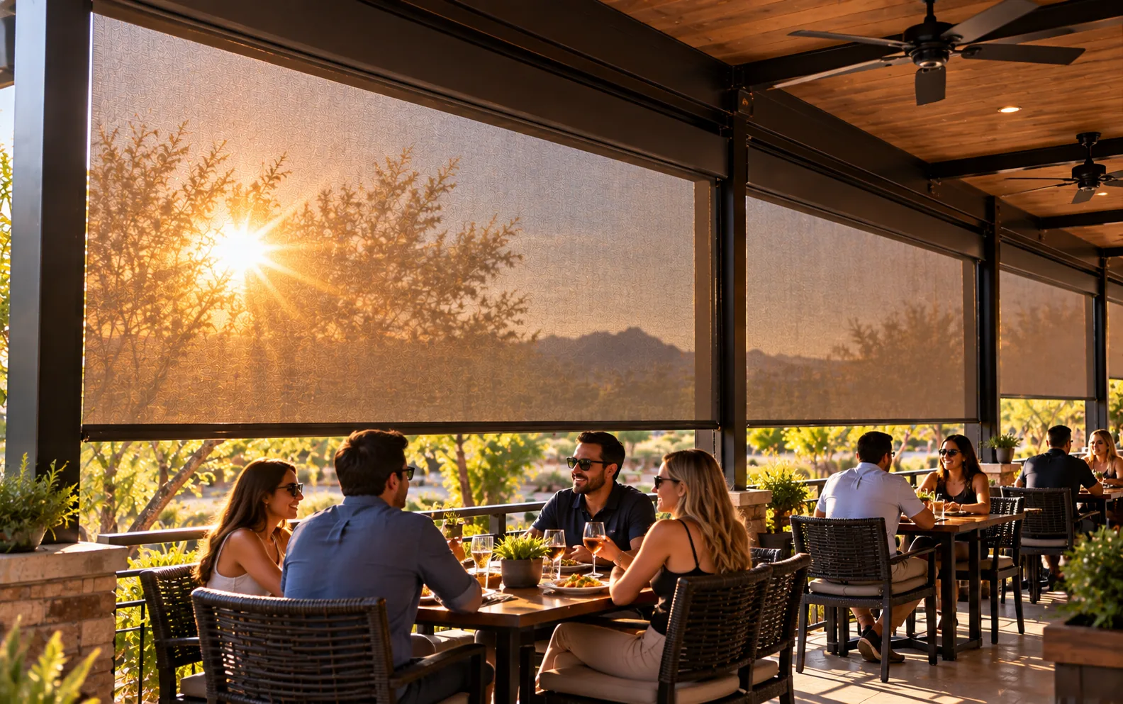 Motorized screens lowered along a covered restaurant patio at sunset, the low sun softened by the mesh and diners seated in the shade below