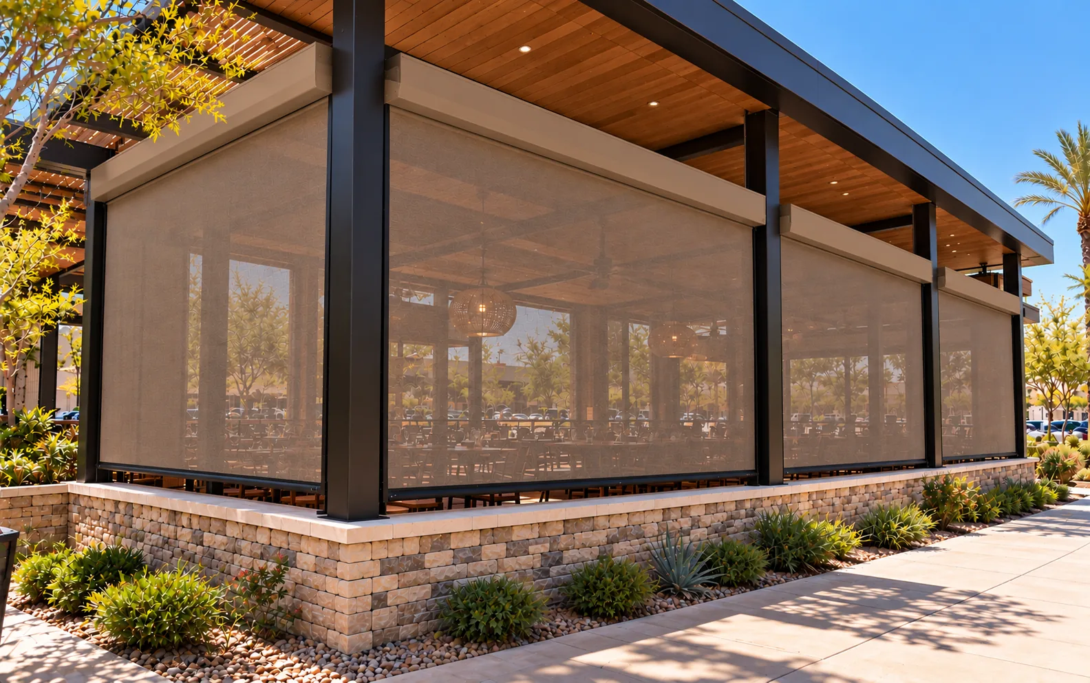 Row of motorized screens lowered across several bays of a covered commercial patio, each screen fitted between the columns above a stone base wall