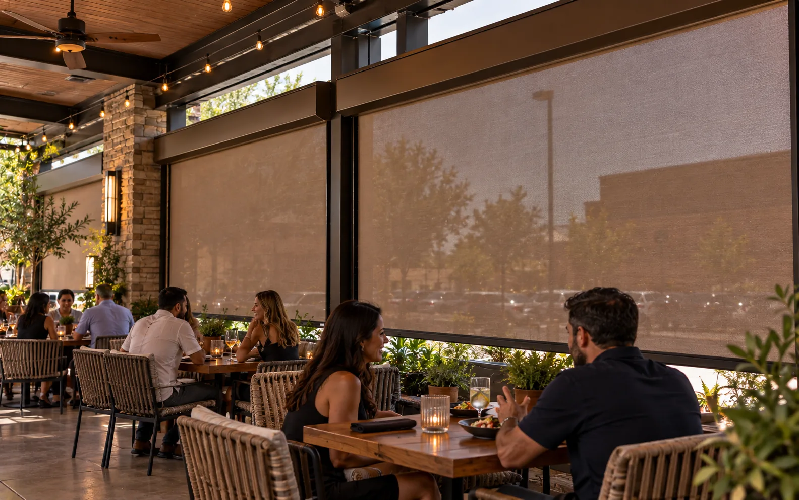 Screens lowered along the parking-lot side of a covered patio, softening the view of cars and a neighboring building behind the dining tables