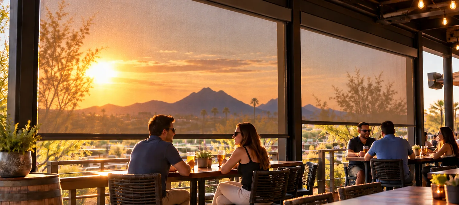 Motorized screens lowered along the rail of a covered bar patio at sunset, with desert mountains and palm trees visible through the mesh and high-top tables below