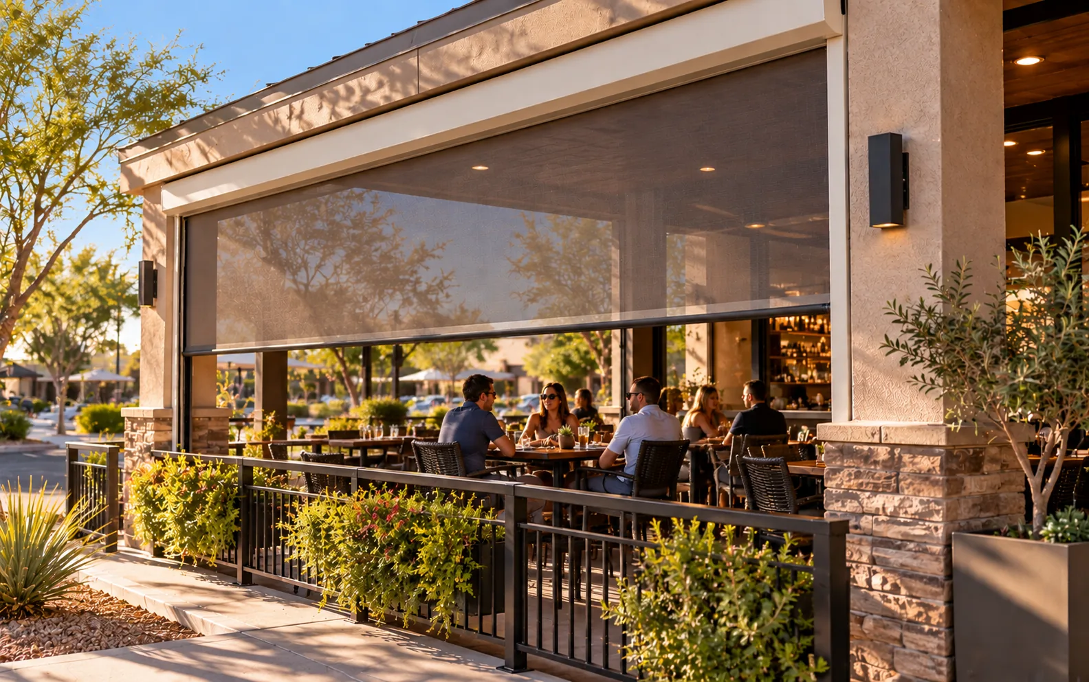Motorized screen partly lowered across the open side of a covered restaurant patio in golden afternoon light, with a stone column, railing planters, and dining tables in the shade below