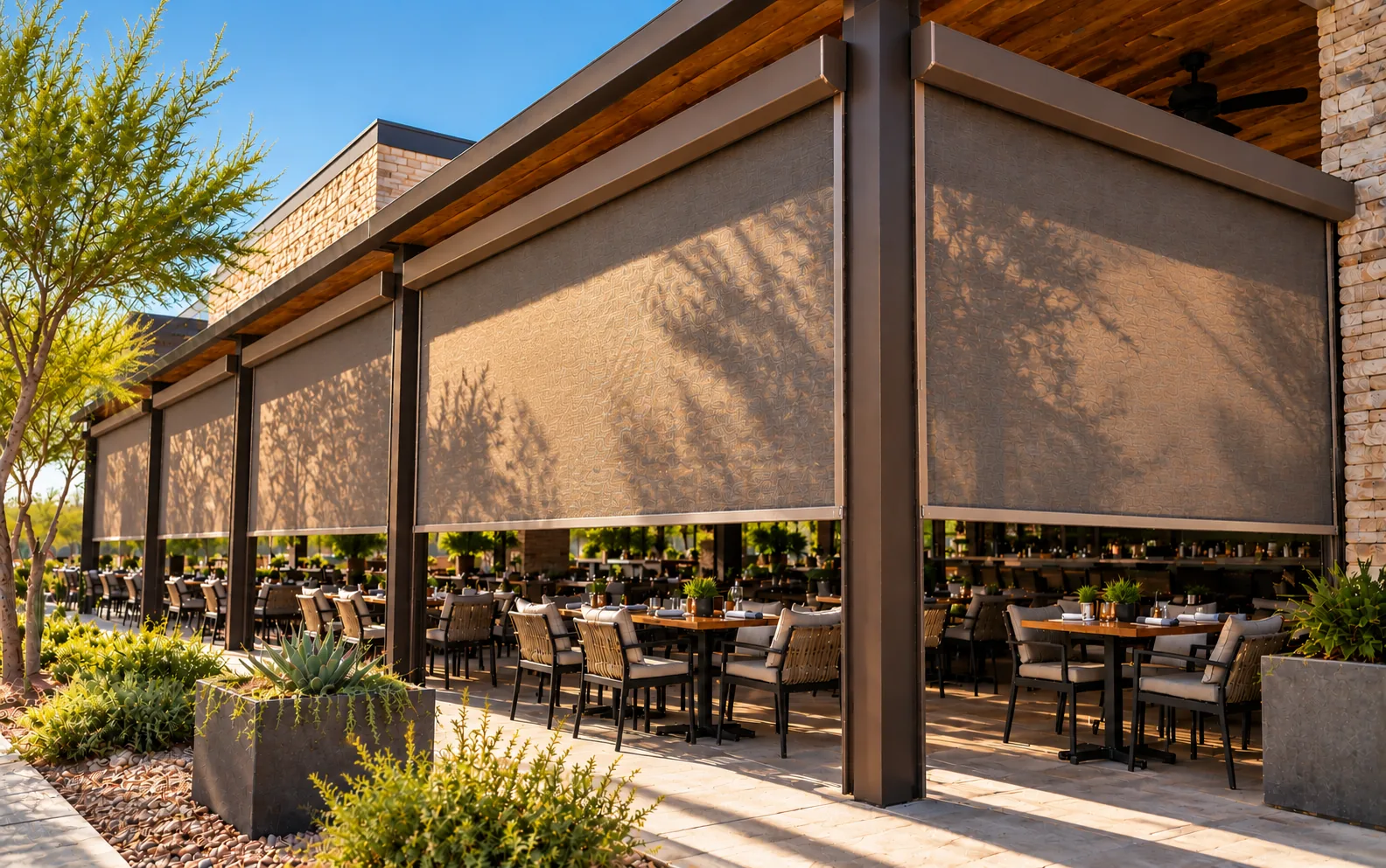 Motorized screens lowered between the columns of a long covered restaurant patio in warm afternoon sun, with tree shadows on the mesh and set dining tables in the shade below