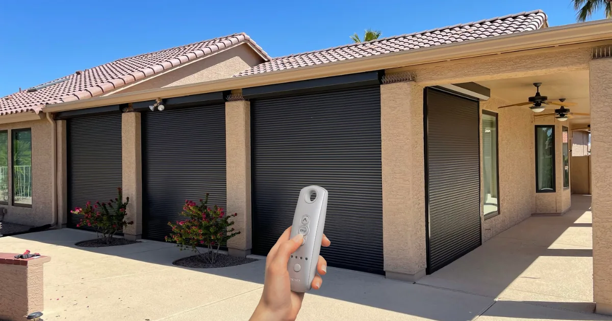 A hand holding a remote in front of a Metro Phoenix home with bronze rolling shutters closed across a row of openings