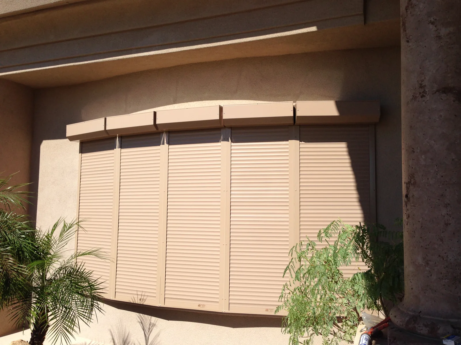 Five tan rolling shutters lowered over a curved bay window on a stucco home, each fitted to its own section of the opening