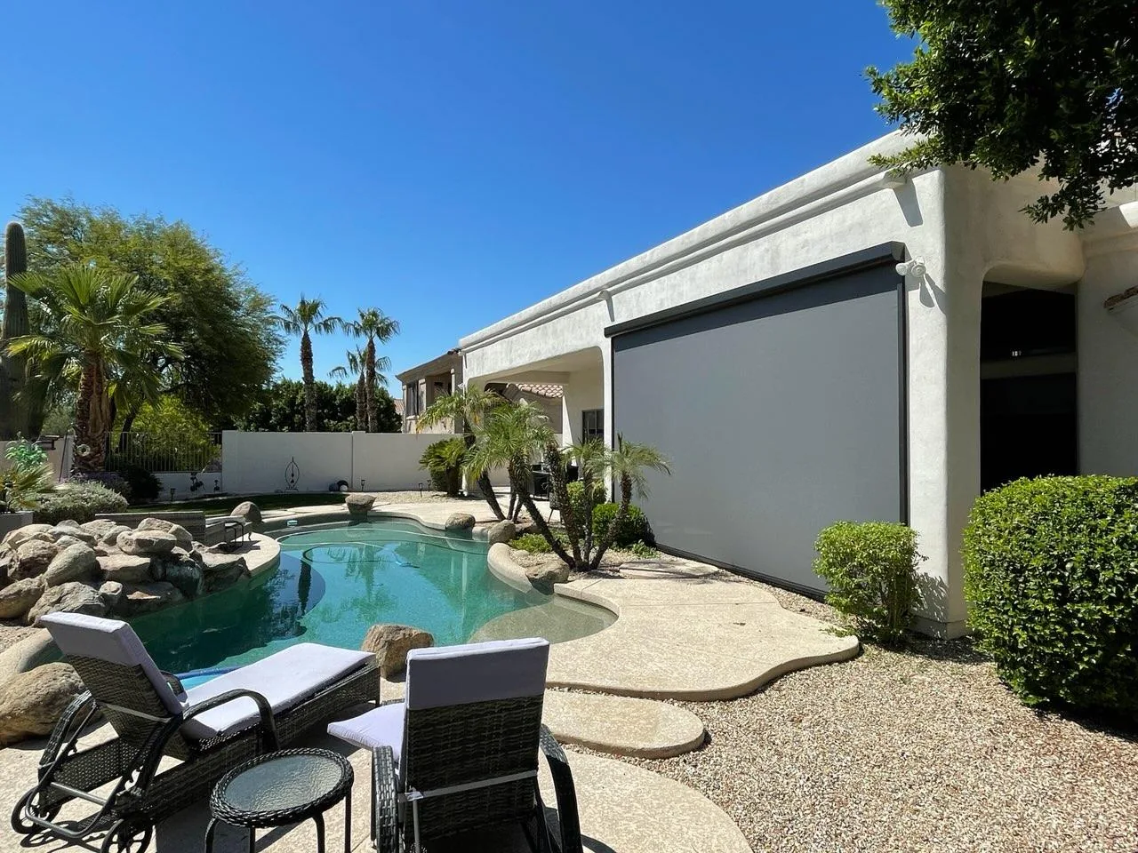 Gray rolling screen lowered across the covered patio of a white stucco home beside a pool with desert landscaping