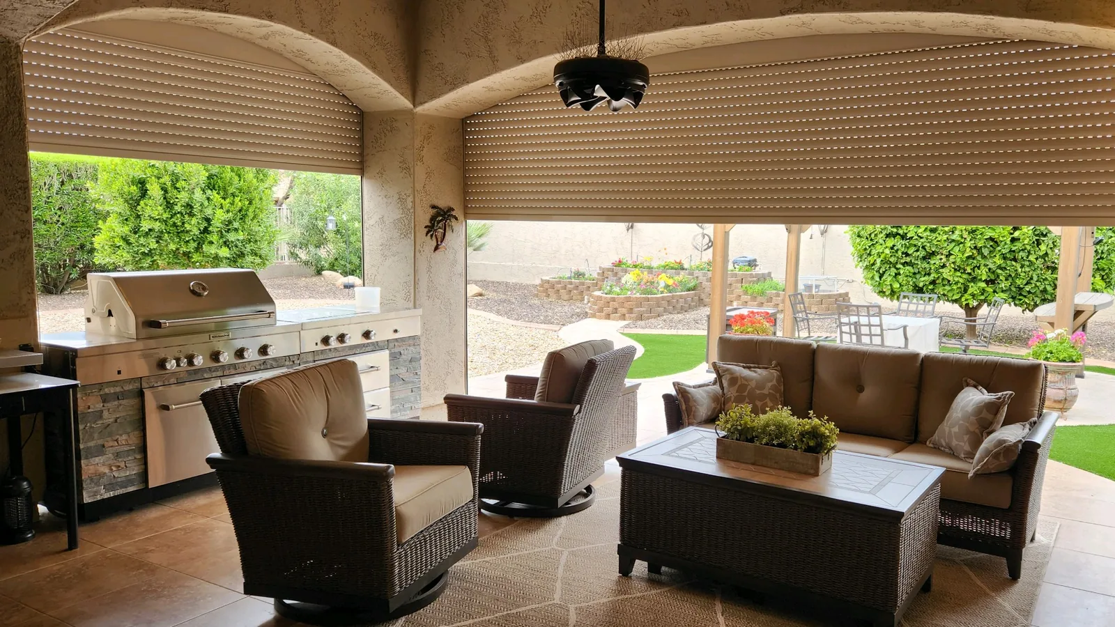 Tan aluminum rolling shutters partly lowered across the arched openings of a covered patio with an outdoor kitchen and seating