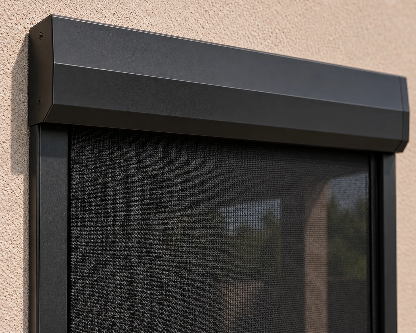 Close-up of a dark powder-coated rolling screen housing above its side tracks, with the mesh lowered on a stucco wall